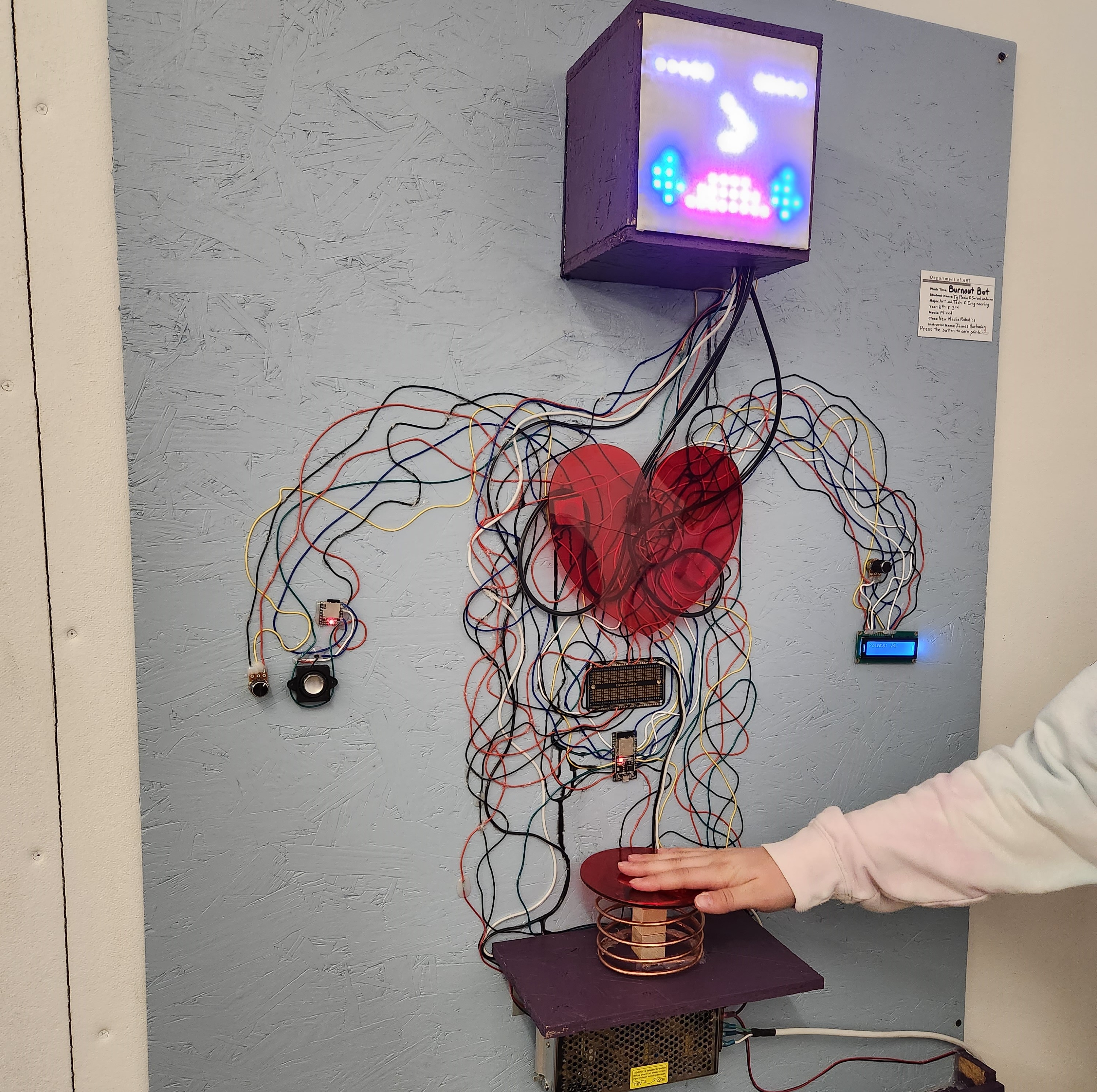 Photo with a hand pressing a large button on the Burnout Bot, which has been installed on a wall. The Burnout Bot is composed of wire art connecting several electronic components, a tubing system carrying oil, and other materials used for aesthetic or construction purposes. In this photo, the Burnout Bot's face screen has a simple crying face animation.